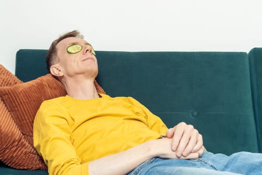 Adult man lying on sofa with eye patches from fresh cucumber slices in living room. Middle aged person reclining on pillow with mask for dark circles under eyes. Home relaxation, beauty treatment.