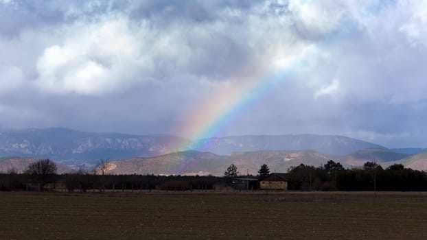 Bright full rainbow on the background of storm cloud over mountain range and valley. Harsh mountain landscape before downpour