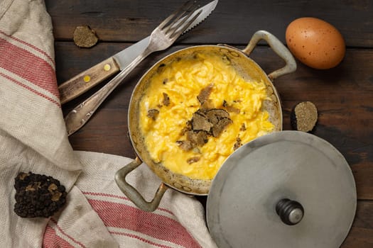 Scrambled eggs with sliced fresh black truffles from Italy served in a a frying pan near fork and knife on wood top view. Italian cousine, gourmet breakfast