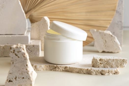 Opened white cream jar with a lid on biege travertine stones against dried palm leaf, close up, mockup. Skincare beauty product. Eco friendly cosmetics
