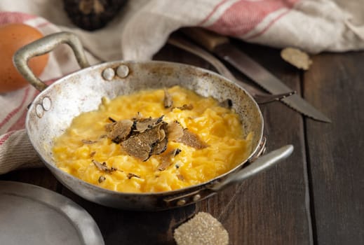 Scrambled eggs with sliced fresh black truffles from Italy served in a a frying pan near fork and knife on wood close up. Italian cousine, gourmet breakfast