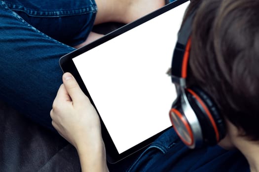 Digital tablet computer with blank white screen in male hands, from above view closeup. Young guy in headphones working on gadget and listen to music. Tablet in hands, touchscreen, electronics.