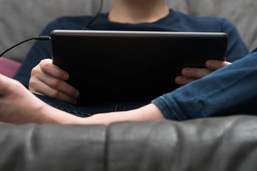 Digital tablet computer in male hands, from below view closeup. Young guy sitting on sofa play videogame on handheld gadget. Modern technology, tablet in hands, touchscreen, electronics.