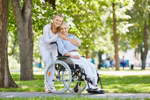 Young female in uniform with senior patient in a wheelchair chilling out in park