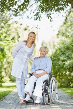 Happy nurse and senior woman in a wheelchair looking at camera in park