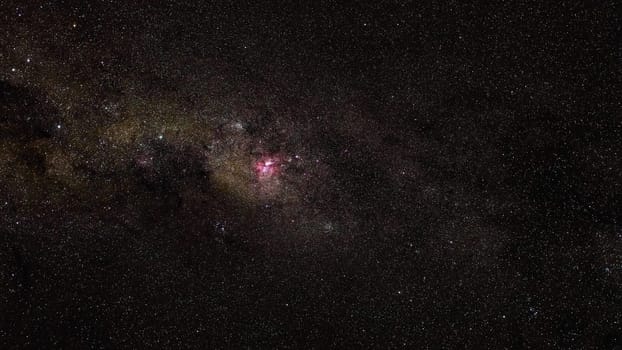 Southern night sky, many stars with milky way around Crux and Vela constellation, red purple Great Carina Nebula in middle visible.