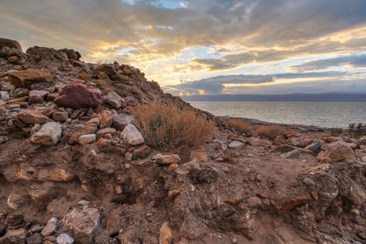 Dead sea shore at Jordan side, dry sand and rocks beach, sunset sky above calm water background.