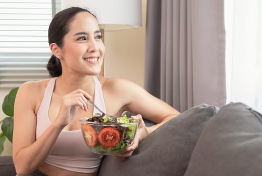Young beauty woman wearing a sportive outfit and enjoy heathy meal with vegetable salad on sofa after yoga execise at home with happiness. Healthy lifestyle and exercise concept