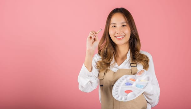 Portrait Asian young woman artist holding brush and paint palette, Happy female painting using paintbrush and palette with colors, studio shot isolated on pink background, Art design workshop