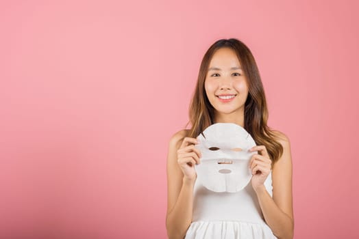 Healthcare spa. Asian woman holding purifying mask face on hands, Portrait beautiful female smiling holds facial sheet mask, Studio shot isolated on blue background, Face care and beauty treatments