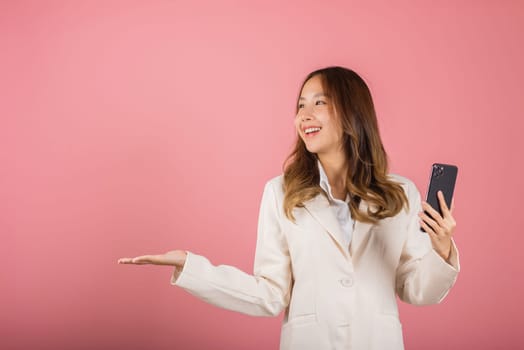 Happy Asian portrait beautiful cute young woman excited holding mobile phone blank screen presenting product with palm of hand at empty, studio shot isolated on pink background, female look to space