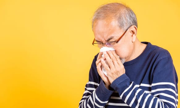 Portrait senior old man blowing nose with tissue studio shot isolated on yellow background, Asian elder man cold having flu and sneezing from sickness virus problem use tissue, pensioner unwell