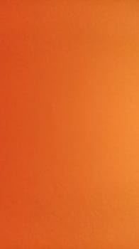 A sheet of thick paper of bright orange color. Close-up. Background intense shade. Natural lateral lighting. Fine cardboard or paper texture. Light gradient in degree of illumination. Vertical image