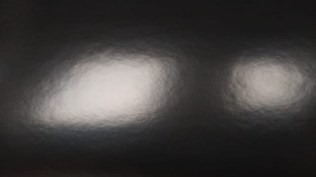 Glare of light on shiny gray paper. Several light spots on a dark surface that reflects light. Abstract monochrome background