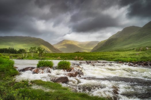 Bundoragha River with small waterfalls and Glenummera and Glencullin mountain ranges illuminated by sunlight, dramatic dark sky, Mayo, Ireland
