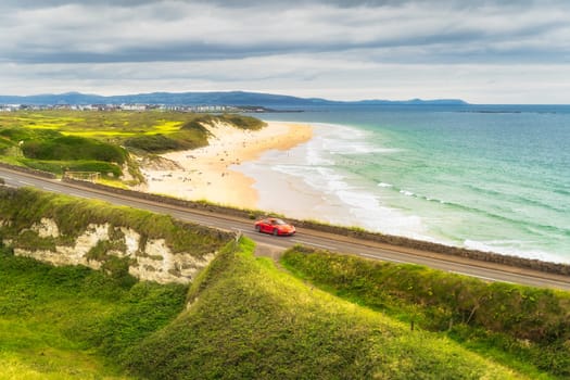 Red sports car on a road with White Rocks Beach and emerald water in background. Causeway Coast and Wild Atlantic Way, Antrim, Northern Ireland