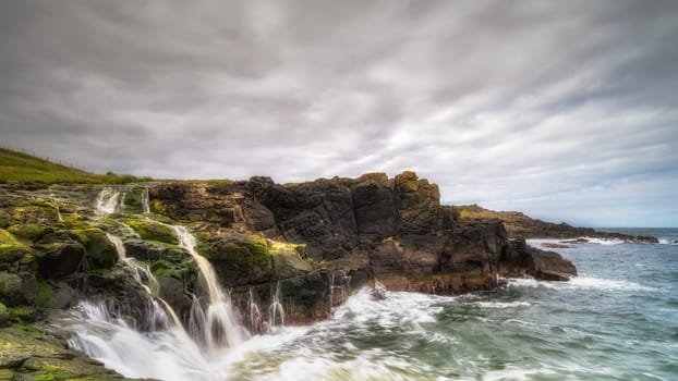 Dunseverick Falls with small cliffs and streaks of water falling down to Atlantic Ocean, Causeway Coast, County Antrim, Northern Ireland