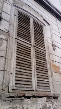 Closed shutters on a high window with platbands. The facade of the building that has not been repaired for a long time. Peeling plaster under the window. The architecture of the old European city.