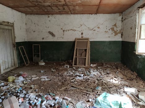 Serbia, Belgrade, May 10, 2020. Inside abandoned house. Grunge scene. Scattered trash on the floor. Green paint on the walls. White half wall. Shot wooden window frames. Bottles, pieces of paper and other items left.