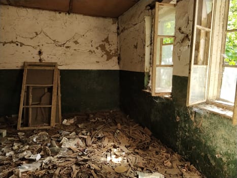 Inside an abandoned house. Scattered trash on the floor. Green paint, cracks and cobwebs on the wall. The white upper half of the wall. Open windows through which sunlight shines through. Frames removed.