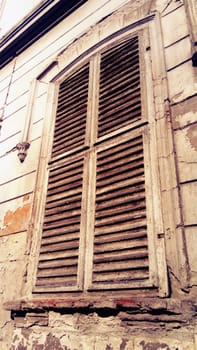Closed shutters on a high window with platbands. The facade of the building that has not been repaired for a long time. Peeling plaster under the window. The architecture of the old European city