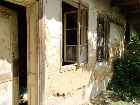 Wall with old windows and a door in a collapsing rural house. The concept of urbanization and oblivion of the motherland. Abandoned house. Broken glass in the windows, open door, falling plaster.