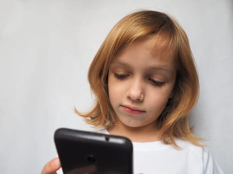 A 7 year old girl with blond hair looks at a smartphone, phone or tablet. Child in white clothes on a gray background. The concept of development of technologies and telecommunications