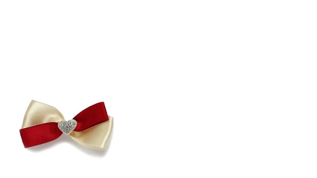 Nice bow made of silky fabric or satin ribbon of beige and red colors. In the middle of the bow there is a heart with shiny silver sparkles. White background. Decoration with light shadow.
