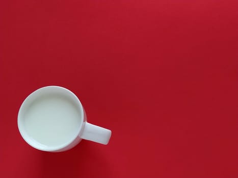 White cup on a red background. In a mug of white milk. View from above. Natural lighting from above and from the side. Flat lay. Greeting card or background. Free space for text. Light ceramic dishes on the left side of the photo.