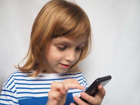 Little girl with mobile phone in her hands. Studio shot. The girl pokes her finger at the smartphone and smiles. Child with blond hair.
