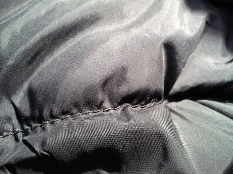 Closeup of black fabric on a winter jacket. Demi-season clothing. Smooth seam with black thread. Slightly shiny synthetic fabric with a seam. Rainproof impregnation. Soft folds. Light and shadow.