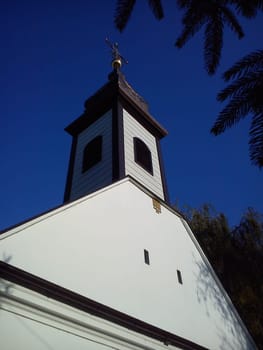 Roof and church dome with a cross. Christian temple, plastered and painted white. The sound of a small wind window and a belfry with a bell. Gilded cross against a blue sky.