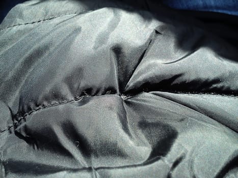 Closeup of black fabric on a winter jacket. Demi-season clothing. Smooth seam with black thread. Slightly shiny synthetic fabric with a seam. Rainproof impregnation. Soft folds. Light and shadow.