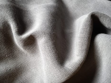 Close-up and texture of a gray knitted fabric, slightly folded in waves or wrinkled. The play of light and shadow on a machine-knitted fabric in natural daylight. Soft, thick, warm cotton material