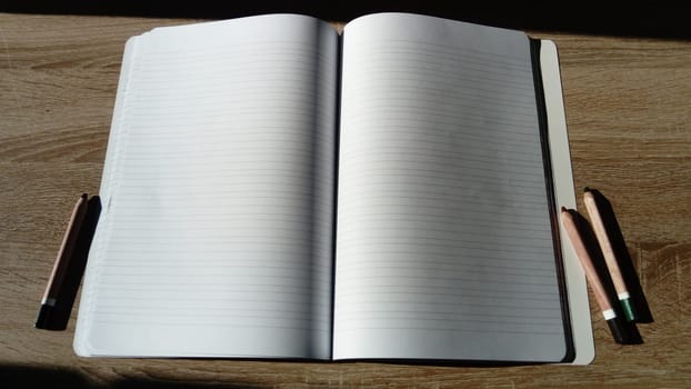 U-turn notebook with blank white pages. To the left and right of the notebook on the wooden table are colored pencils. Harsh natural sunlight