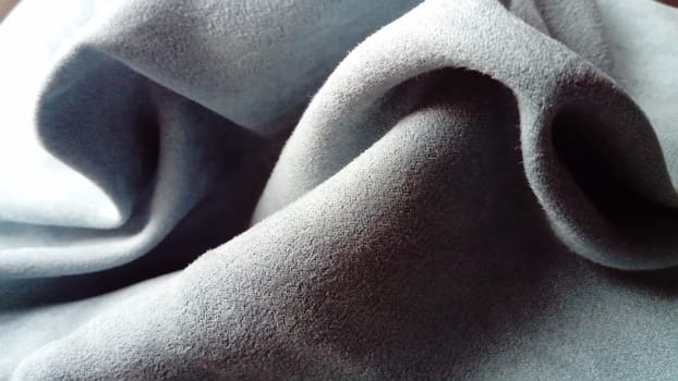 A close-up of a velvety dense fabric for curtains of gray-blue color, randomly folded by beautiful smooth waves with a play of light and shadow. Natural daylight