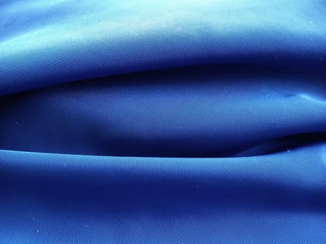 Synthetic fabric in blue, photographed at close range. A noticeable interweaving of threads. Fabric with anti-rain and anti-wind impregnation. A piece of material is laid in waves, light and shadow.