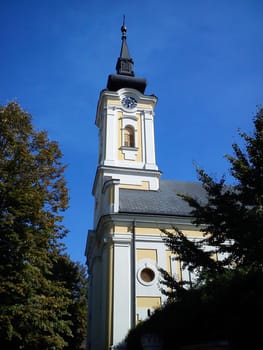 Part of the church building with a dome and a clock under it. The temple is painted in white - yellow colors, the dome and spire - black. Early autumn with blue sky.