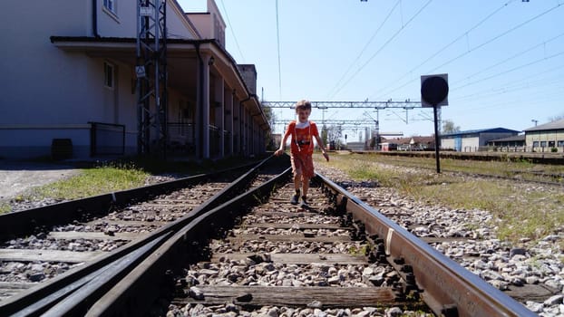 Sremska Mitrovica, Serbia, June 1, 2020. Boy with a protective mask runs on the railroad. Railroad tracks and station. Danger on the rails. Protection against respiratory viral diseases.