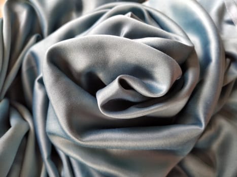 Silky to the touch fabric of gray - blue color with a brilliant shimmer. Natural lighting, light and shadow. The material is casually folded into a circle, resembling a rose bud. Polyester Textile.