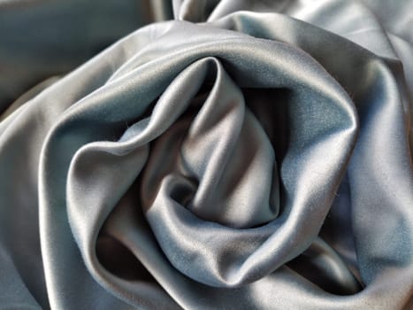 Silky to the touch fabric of gray - blue color with a brilliant shimmer. Natural lighting, light and shadow. The material is casually folded into a circle, resembling a rose bud. Polyester Textile