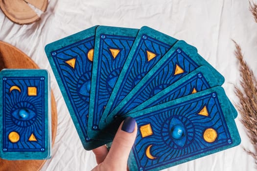 Hand with blue nails hold deck of Tarot cards fanned out over white cloth sheet with wooden tray and spikelets. Top view. Minsk, Belarus - 09.27.2021