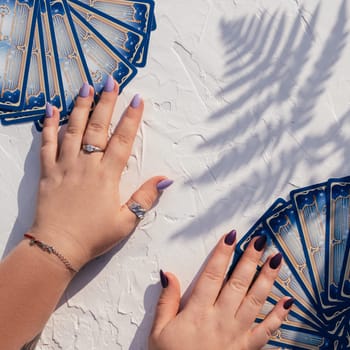 Hands with purple nails and rings on deck of Tarot cards fanned out on white surface with fern leaf shadow. Top view. Minsk, Belarus - 07.28.2021