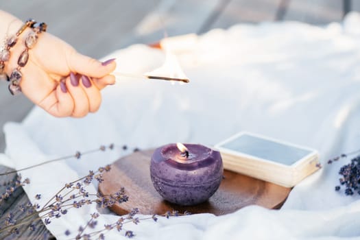 Woman's hand in bracelet with purple long nails lights candle with match, next to deck of cards and lavender flowers on white and wooden surface