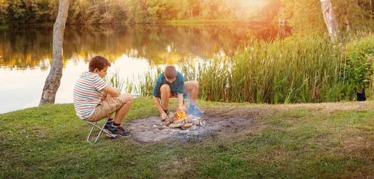 Two teenagers lighting a campfire in nature near the lake. Concept of the scout's camping and family vacation.