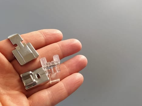 A set of feet for an electric sewing machine in a female hand. Gray backdrop. Standard sewing or zigzag foot, for sewing in zippers, overlock foot and for buttons.