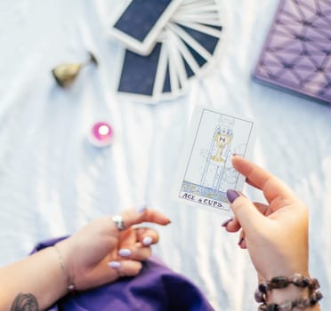 Female hand with purple nails holds tarot card named Ace of Cups over white surface with notebook and candle. Top view. Minsk, Belarus - 07.28.2021