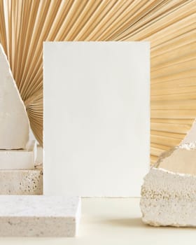 Blank card near beige travertine stones against dry beige palm leaf close up, mockup. Boho scene with vertical paper card. Bohemian or close to nature greeting or wedding Invitation. Minimal composition.
