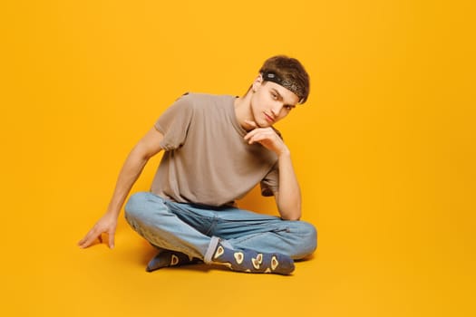 A young adult man dressed in rolled jeans and olive t shirt, sitting against a vibrant yellow studio background