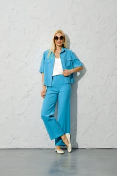 Fashion model in sunglasses in blue jacket and trousers posing leaning against textured white wall